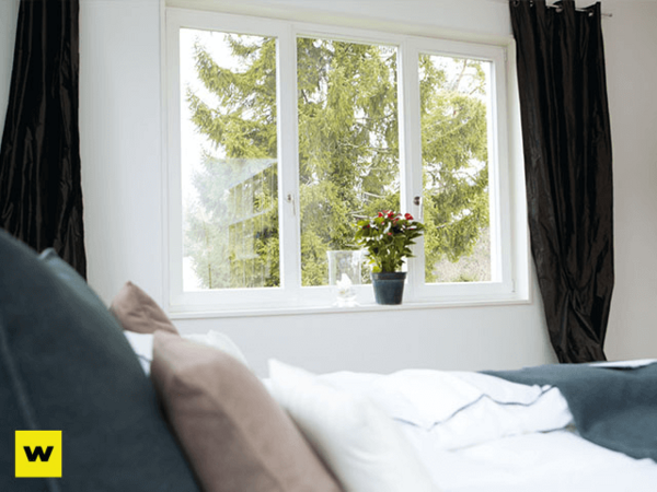 Modernes Schlafzimmer mit großen Fenstern, Blick ins Grüne und Zimmerpflanze auf der Fensterbank.