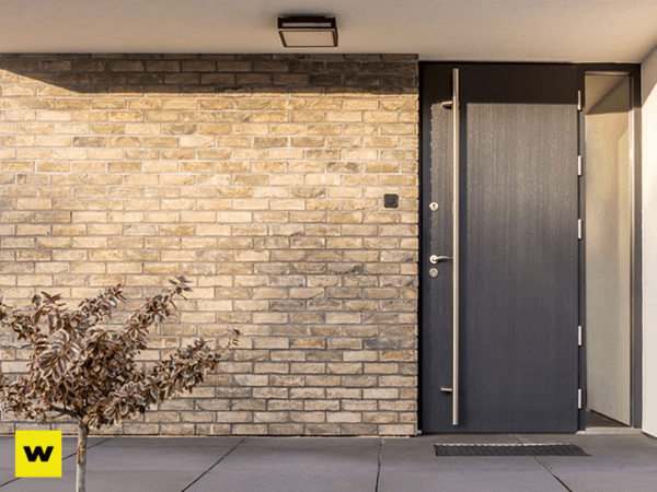 Moderne Haustür aus dunklem Holz vor heller Klinkerfassade mit kleinem Baum im Vorgarten.